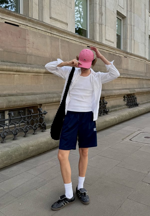 Person steht auf einem Bürgersteig in der Stadt und trägt eine rosa Kappe, ein weißes Hemd und Tanktop, marineblaue Shorts, weiße Socken, schwarze Schuhe und trägt eine schwarze Tasche.