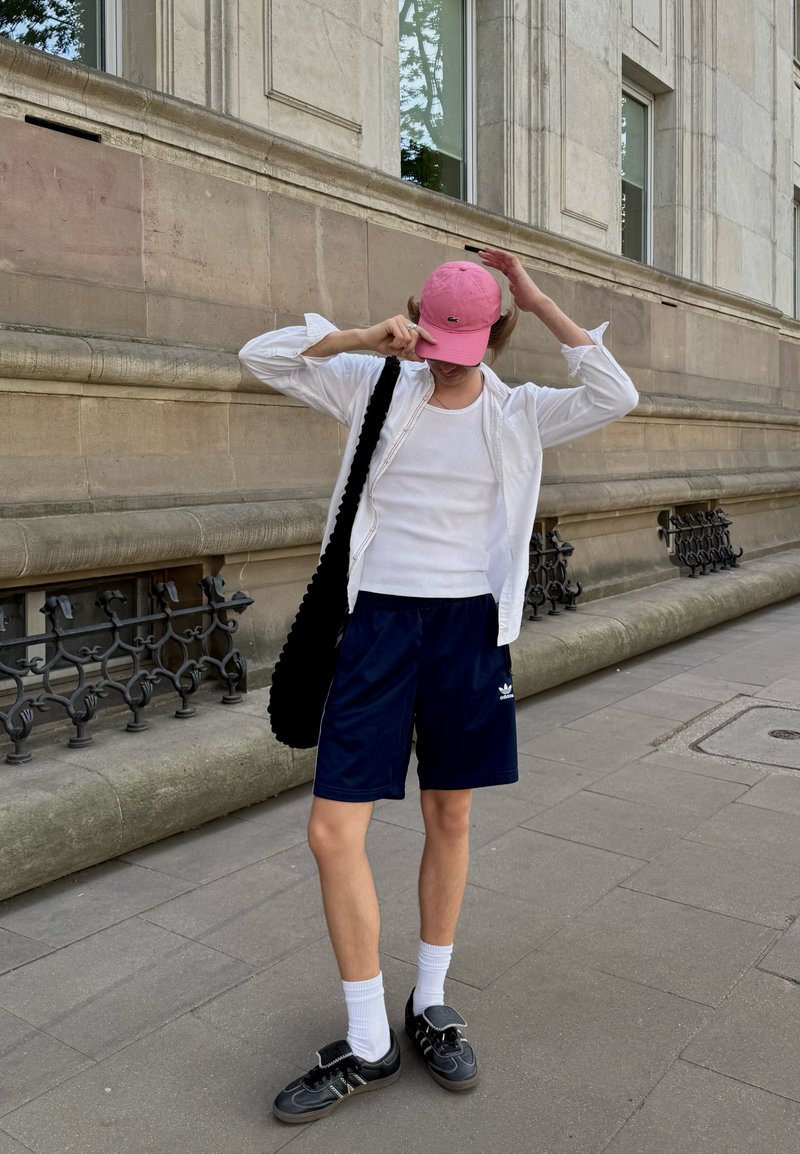 Personne debout sur un trottoir en ville portant une casquette rose, une chemise blanche et un débardeur, un short bleu marine, des chaussettes blanches, des chaussures noires, et portant un sac noir.