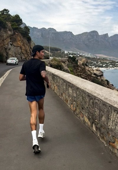 Hombre corriendo por carretera costera con muro de piedra, acantilados rocosos y montañas bajo un cielo nublado, coche acercándose por detrás.