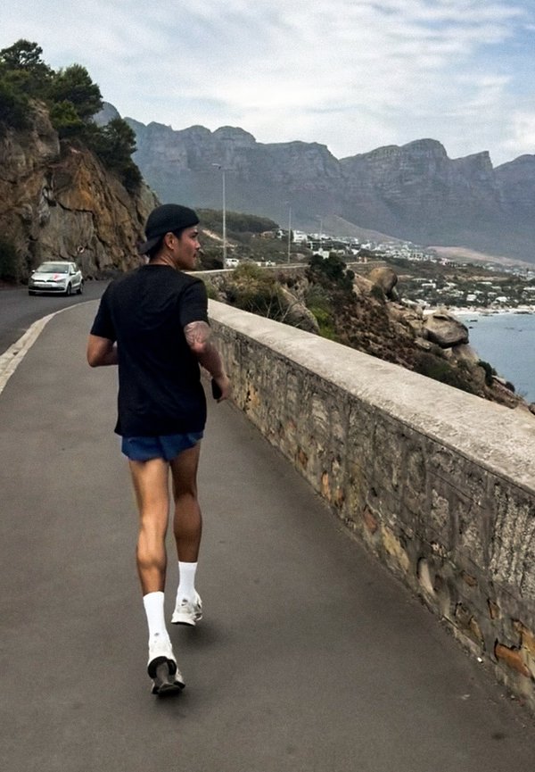 Homme jogging sur une route côtière avec un mur en pierre, des falaises rocheuses et des montagnes sous un ciel nuageux, voiture approchant par l'arrière.