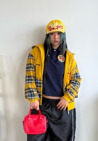 Gul og rutete jakke, marineblå polo, svarte bukser, holder en liten rød veske med to håndtak og logo. Langt mørkt hår med blå striper.