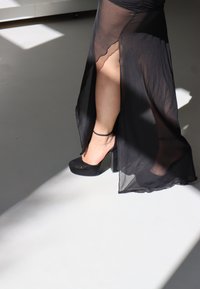 Saia maxi preta transparente com fenda lateral, combinada com sapatos plataforma de salto alto pretos. Apresenta uma textura suave e detalhe de tiras no tornozelo.
