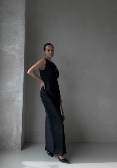 Vestido negro sin mangas con un corpiño ajustado y una falda fluida, combinado con tacones negros de punta y un pequeño bolso. Tela suave contra un fondo de concreto.