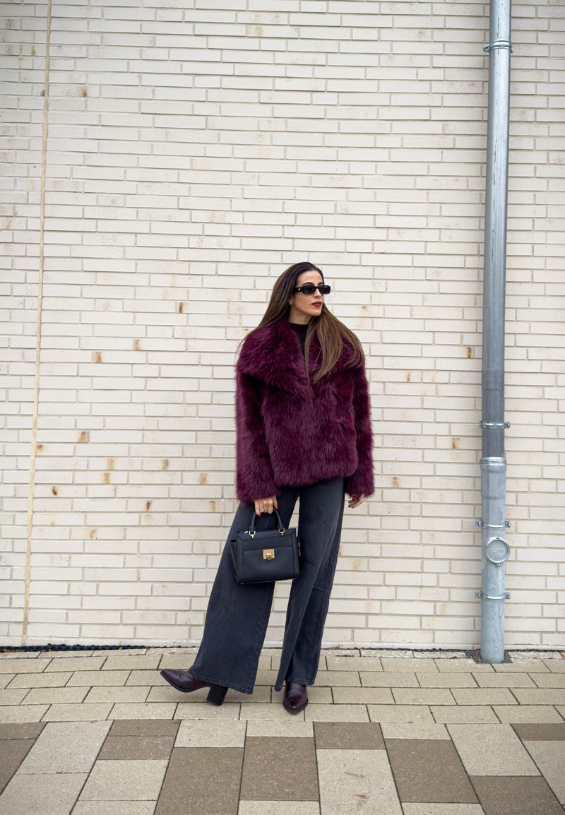 Vinröd jacka i konstpäls med en bred krage, kombinerad med svarta vida byxor och en liten svart handväska. Modellen bär svarta solglasögon.