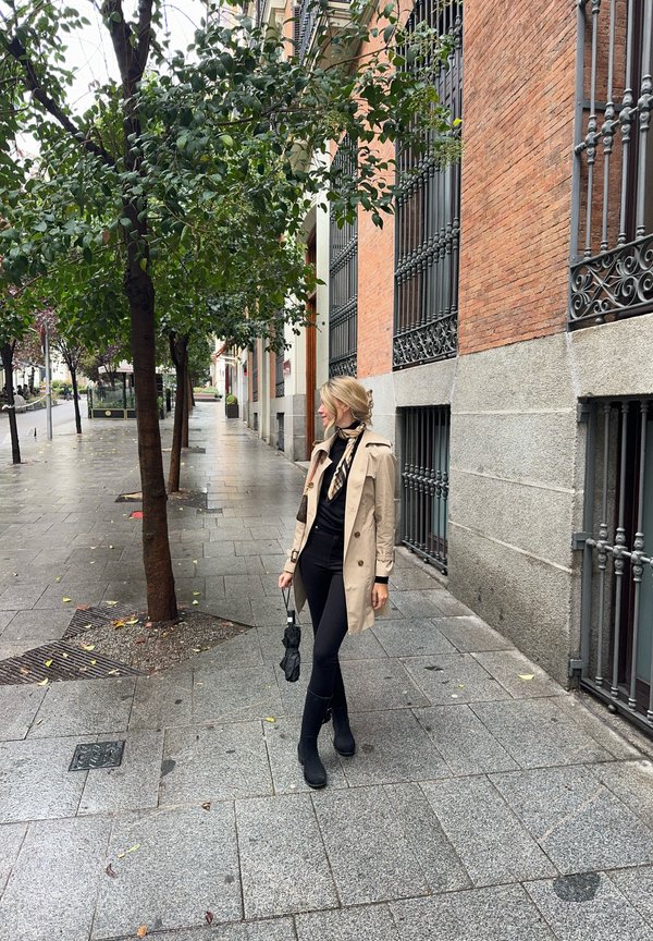 Abrigo trench beige sobre una camiseta y pantalones negros, sosteniendo un pequeño paraguas negro, de pie sobre un pavimento mojado con edificios de ladrillo y árboles cerca.