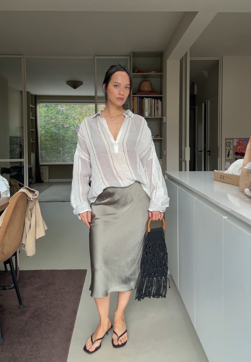 Chemise en lin rayée légère avec un col à boutons, associée à une jupe satinée vert olive. Tient un sac en macramé noir, porte des tongs.