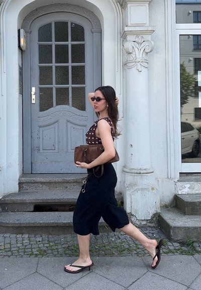 Mujer con gafas de sol y camiseta de lunares caminando descalza en chanclas frente a una puerta gris en una calle adoquinada.