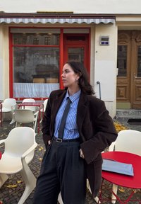 Hnedé kordurové sako, modro-prúžkovaná košeľa na gombíky, sivé plisované nohavice a čierna kravata. Mestské kaviarne s bielymi stoličkami a červeným stolom.