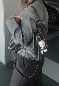 Ženska nosi sivo blazer jakno in črne hlače, nosi črno usnjeno torbico z okrasnimi studnimi detajli ter pritrjenim majhnim belim Hello Kitty ključem.
