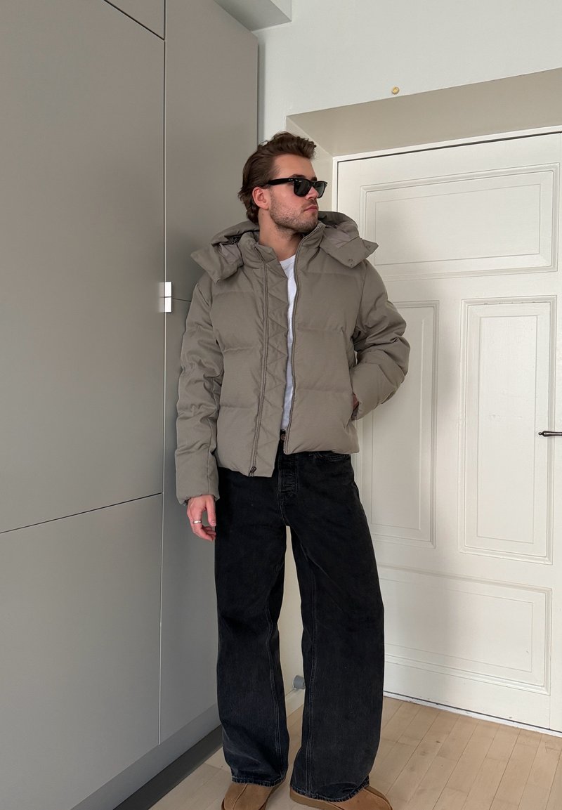 Homme portant des lunettes de soleil, une doudoune beige, une chemise blanche, un jean noir ample et des chaussures marron, se tenant à l'intérieur près d'une porte blanche et de placards gris.