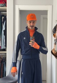 Sportanzug in Marineblau mit weißen Streifen, Reißverschlussjacke, orangefarbener Langarmshirt darunter und orangene Mütze. Hält ein Handy mit buntem Case im Spiegel.