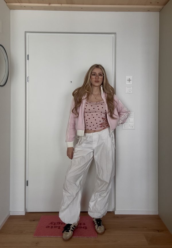 Mladá žena s dlhými blond vlasmi, ktorá nosí okuliare, ružový top s bodkami, svetloružovú bundu, biele voľné nohavice a tenisky, stojí vo vnútri.