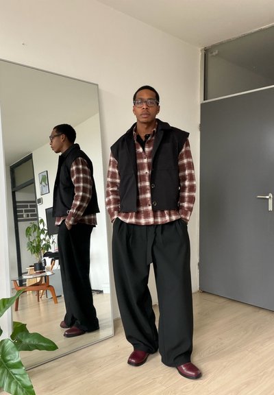 Hombre con gafas, camisa de cuadros, chaleco negro, pantalones anchos negros y zapatos burdeos de pie en el interior cerca de un espejo alto que refleja su perfil lateral.
