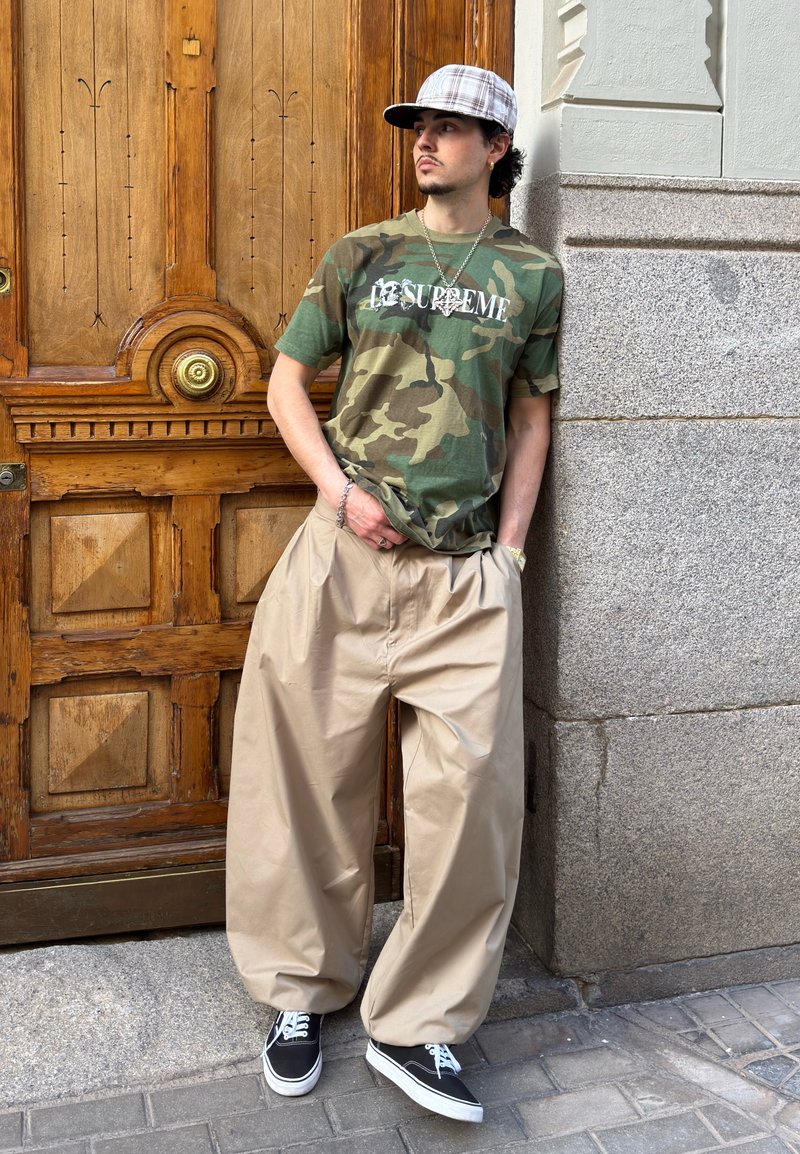 Jeune homme portant un t-shirt camouflage, un pantalon large beige, une casquette à carreaux et des baskets, appuyé contre une porte en bois et un mur en pierre à l'extérieur.