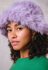 Chapéu roxo fofinho com uma aba larga, usado por uma pessoa com cabelo encaracolado. Um suéter verde texturizado e brincos verdes completam o visual.