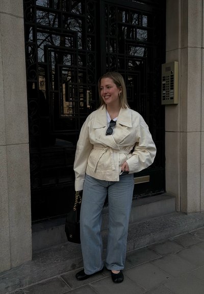 Joven mujer sonriendo, vestida con una chaqueta crema con cinturón, vaqueros azul claro, zapatos planos negros, sosteniendo un bolso negro, de pie junto a una puerta oscura y ornamentada.