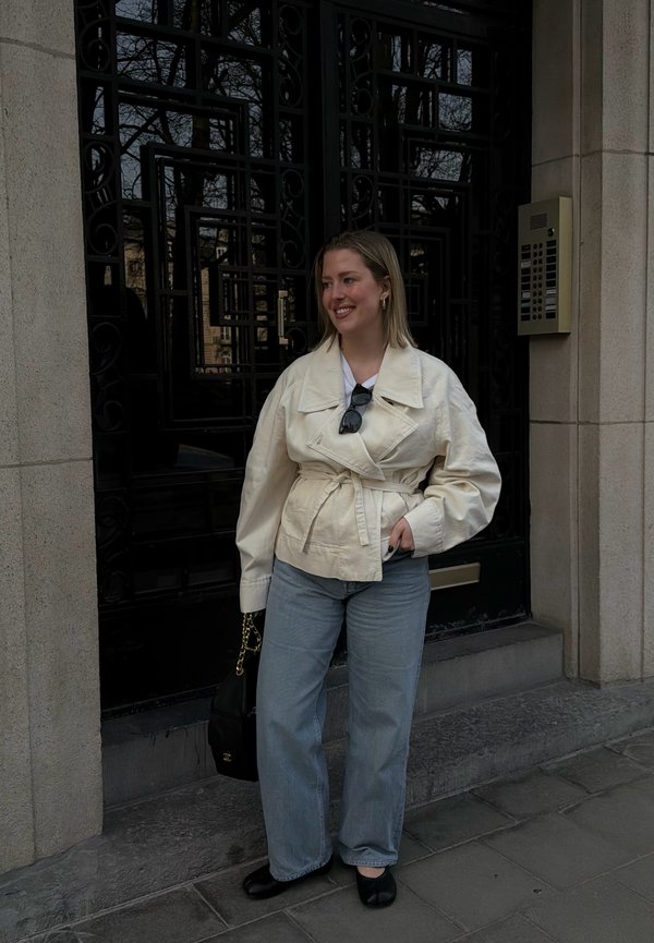 Jeune femme souriante, portant une veste crème ceinturée, un jean bleu clair, des ballerines noires, tenant un sac à main noir, debout à côté d'une porte ornée sombre.