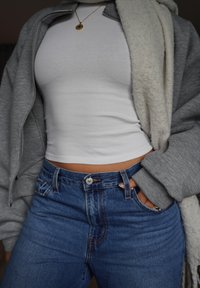 Grå zip-up sweatshirt över en vit linne, kombinerad med höga blå jeans. Noterbara detaljer inkluderar ett halsband med guldfärgad medaljong.