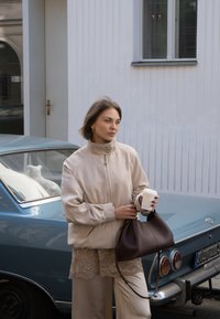 Giacca softshell beige con colletto alto, top in pizzo a strati, pantaloni crema e borsa in pelle marrone. Tiene in mano una tazza di caffè, capelli corti castani.