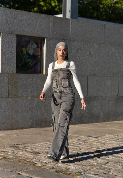 Pantalones de mezclilla grises con bolsillos y una cremallera, combinados con una camiseta blanca de manga larga. La modelo camina por un camino de adoquines bajo la luz natural.