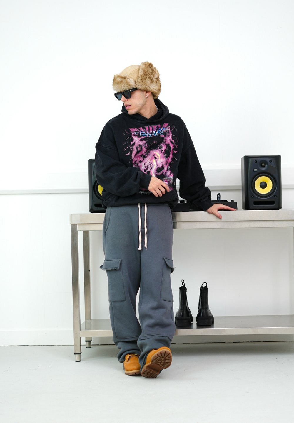 Μαύρη φούτερ με γραφικό σχέδιο, γκρι φόρμες με τσέπες cargo, καφετί μπότες και ψεύτικο γούνινο καπέλο, που στέκεται κοντά σε ηχεία πάνω σε μεταλλικό τραπέζι.