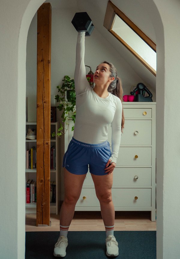 Vrouw in sportkleding die een halter boven haar hoofd tilt in een heldere kamer met een dressoir en kamerplanten.