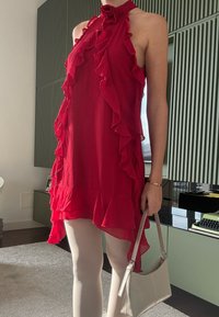 Rotes Neckholderkleid mit Rüschenkanten und gestuftem Stoff. Kombiniert mit cremefarbenen Strumpfhosen und einer hellgrauen Handtasche. Modernes, minimalistisches Design.