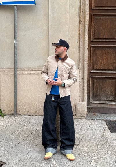 Hombre con chaqueta beige, suéter azul, vaqueros anchos oscuros, zapatos amarillos y gorra negra de pie junto a un muro de piedra y una puerta de madera en la acera.