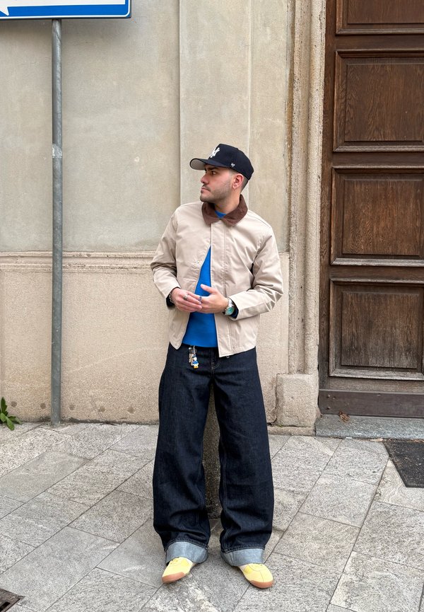 Man wearing beige jacket, blue sweater, wide dark jeans, yellow shoes, and black cap standing by stone wall and wooden door on sidewalk.