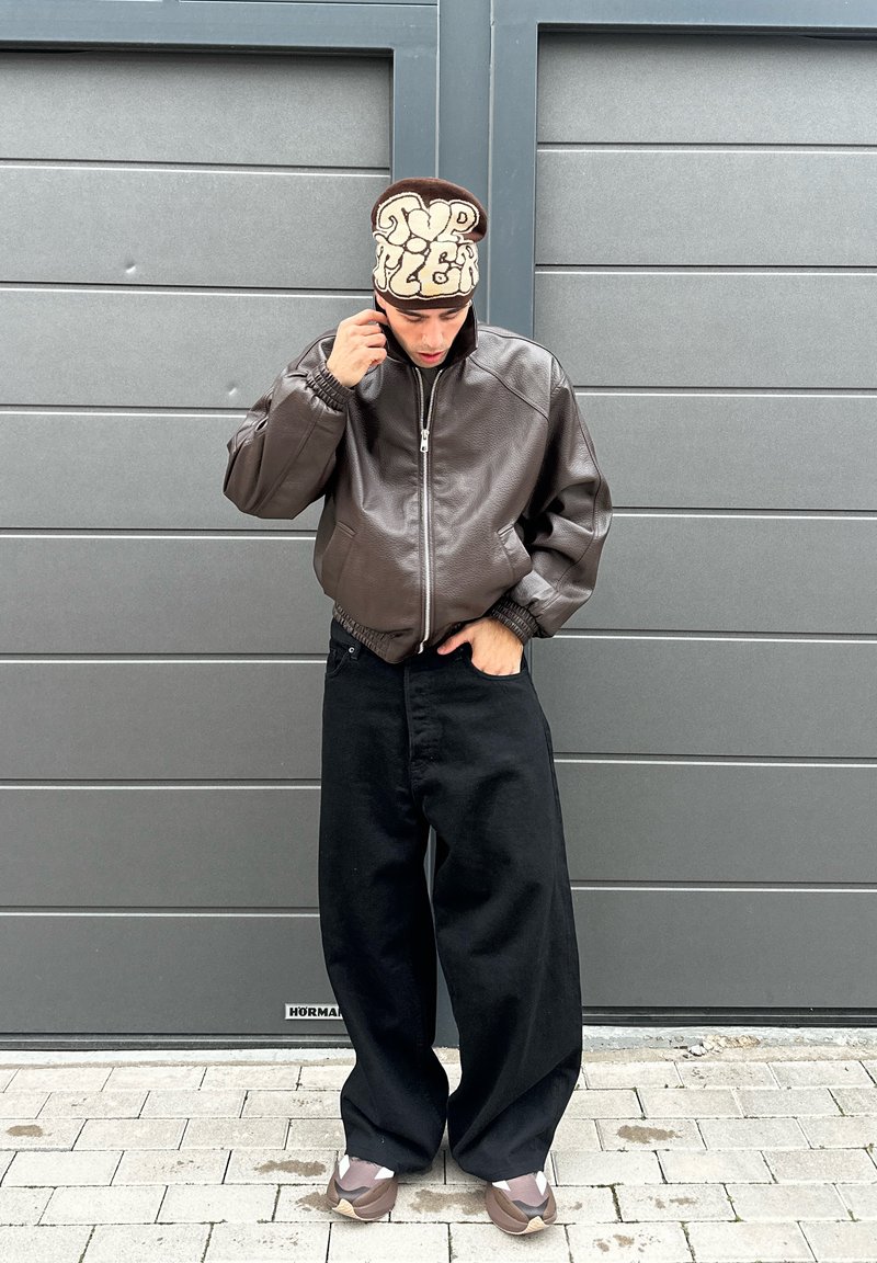 Persona che indossa una giacca di pelle marrone, pantaloni larghi neri e un berretto marrone e beige, in piedi davanti a un portone del garage grigio.