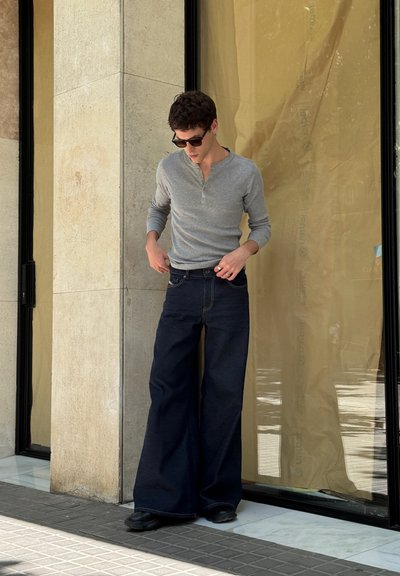 Camisa gris de manga larga y jeans oscuros de pierna ancha. Los detalles destacados incluyen costuras visibles y un ajuste relajado. Unos zapatos negros completan el look.
