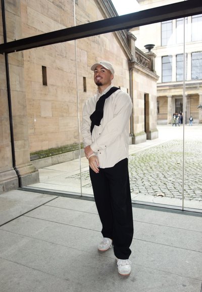 Camisa de botón color beige claro, bufanda negra texturizada sobre los hombros, pantalones negros, zapatillas blancas con suelas marrón claro, en un entorno interior.