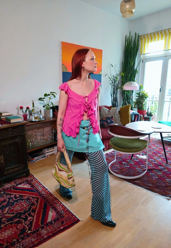 Donna con capelli rossi, con top rosa e pantaloni a righe, che tiene in mano una borsa, in piedi in un soggiorno colorato con piante e tappeti a motivo.