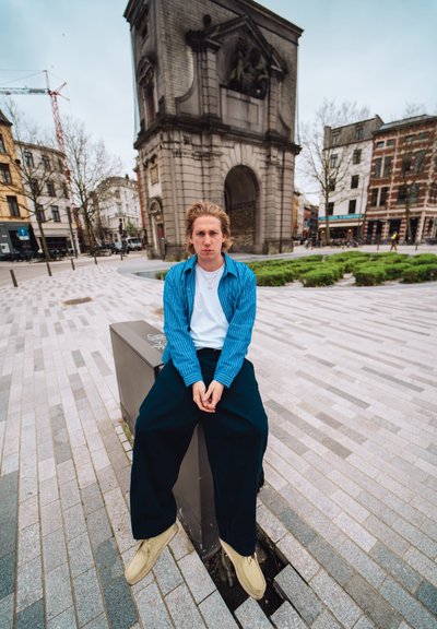 Hombre sentado en un bloque de concreto, vistiendo una camisa de rayas azul, una camiseta blanca, pantalones oscuros anchos y zapatos beige en un entorno urbano.