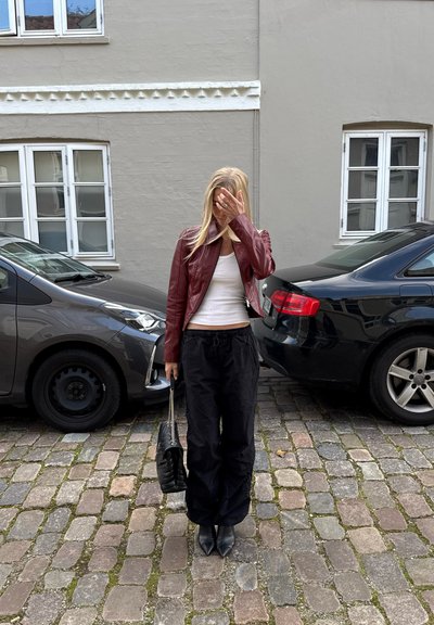 Chaqueta de cuero roja, camiseta blanca sin mangas, pantalones negros holgados y botas negras puntiagudas. Suelo de adoquines, edificio gris, coches estacionados al fondo.