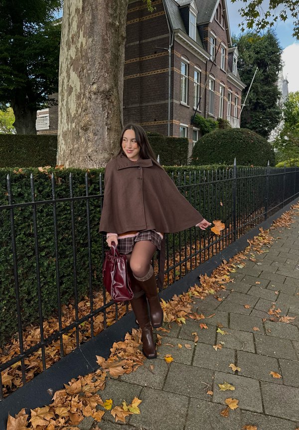 Une femme portant une cape brune, une jupe à carreaux et des bottes montant jusqu’aux genoux tient un sac bordeaux près d’une clôture bordée d’arbres, sur un trottoir jonché de feuilles.