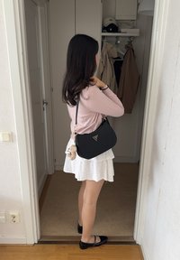 Schwarze Leder-Schultertasche mit goldenen Akzenten, weißer Rock mit Blumenmuster und flachen schwarzen Schuhen, stehen in einer Türöffnung.