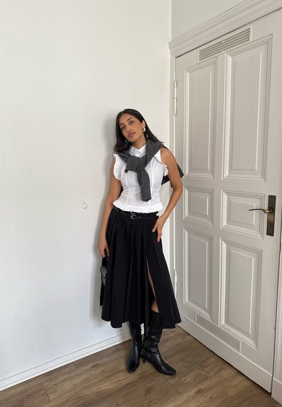 Camisa blanca con volantes, suéter gris atado alrededor de los hombros, falda negra plisada con abertura, botas negras hasta la rodilla, de pie junto a una puerta blanca.