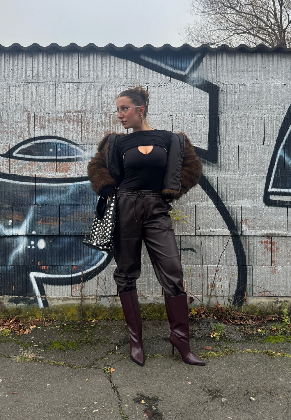 Μαύρο κορμάκι με κενό, καφέ ψεύτικη γούνα, καφέ δερμάτινα παντελόνια και μπορντό τακούνια με μαύρη τσάντα με λευκές βούλες.