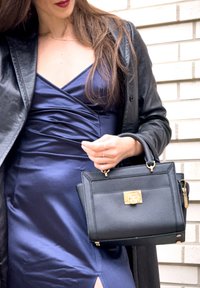 Femme portant une robe satinée bleu marine et une veste en cuir noire, tenant un sac à main structuré noir avec une fermeture dorée, devant un mur en briques blanches.