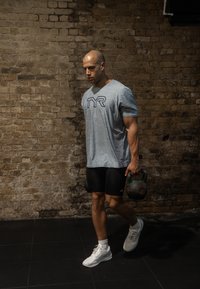 Mann in Sportbekleidung geht drinnen auf schwarzem Boden, hält in der rechten Hand eine Kettlebell, vor einem strukturierten Ziegelwand-Hintergrund.