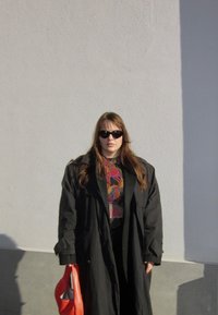 Melns, pārmērīgi liels trenčkotis, krāsains rakstains tops, melnas bikses, tur sarkanu somu, saulesbrilles, stāv uz vienkāršas baltas sienas.