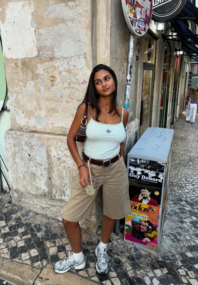 Joven mujer con pantalones cortos y camiseta sin mangas sosteniendo una bebida, apoyada contra una pared de piedra en una calle empedrada con carteles y tiendas cercanas.