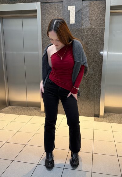 Joven mujer con top burdeos de un solo hombro y pantalones negros está entre dos puertas de ascensor bajo un cartel con el número 1 en un pasillo con baldosas.