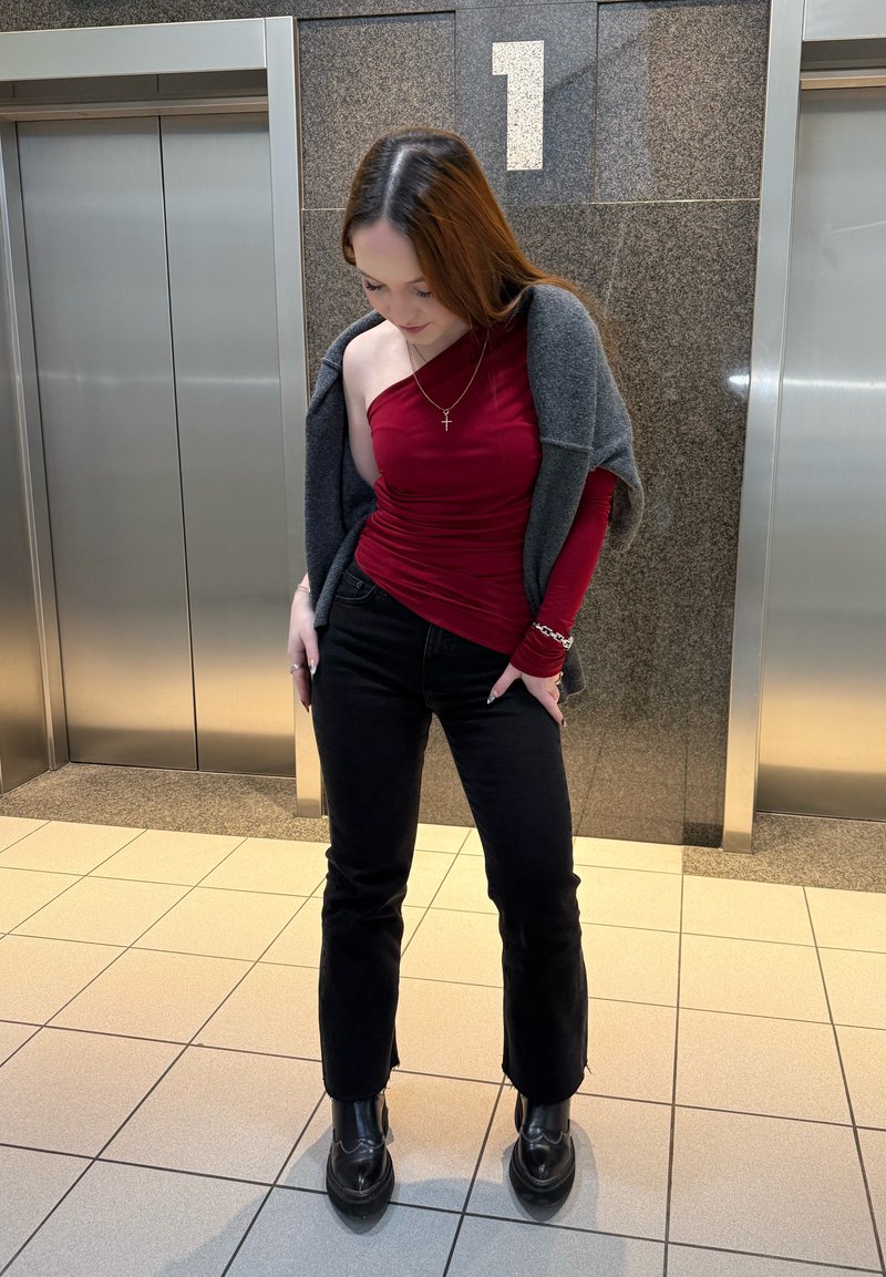 Jeune femme portant un haut bordeaux à une épaule et un pantalon noir se tient entre deux portes d'ascenseur sous un panneau numéro 1 dans un couloir carrelé.