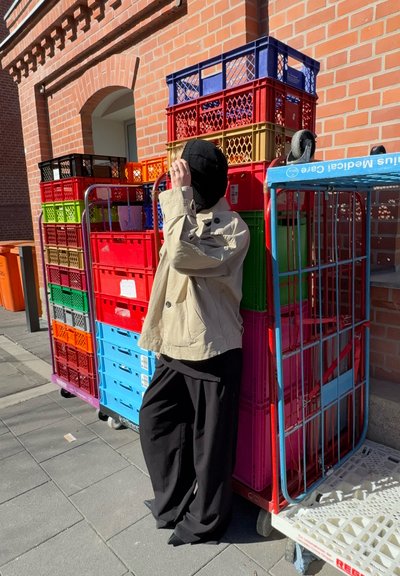 Chaqueta beige, pantalones negros y sombrero, apoyándose en coloridas cajas de plástico apiladas en un entorno urbano de ladrillo rojo con pavimento gris.