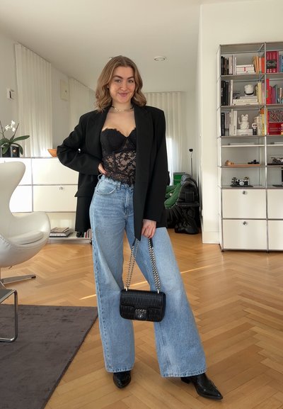 Mujer joven de pie en el interior, vistiendo una blusa negra de encaje, un blazer negro oversize, jeans de pierna ancha, botas negras y sosteniendo un bolso negro.