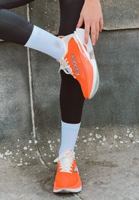 Ljust orange löparskor med vita accenter, med en slät syntetisk ovandel, snören och en dämpad sula, parat med svarta leggings och blåa strumpor.
