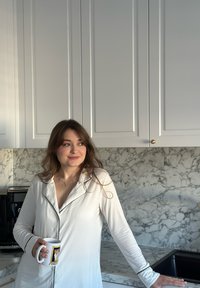 Camicia da notte bianca a maniche lunghe con bordi neri, in piedi accanto a un piano di marmo e un lavandino nero, mentre tiene una tazza bianca con un design di corgi.