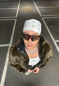 Ψεύτικο γούνινο μπουφάν με μαύρη δερμάτινη λεπτομέρεια, λευκή μαντήλα με σχέδιο, υπερμεγέθη γυαλιά ηλίου με κόκκινες λεπτομέρειες, επάργυρες αλυσίδες σε στρώσεις, λευκή μπλούζα χωρίς μανίκια.